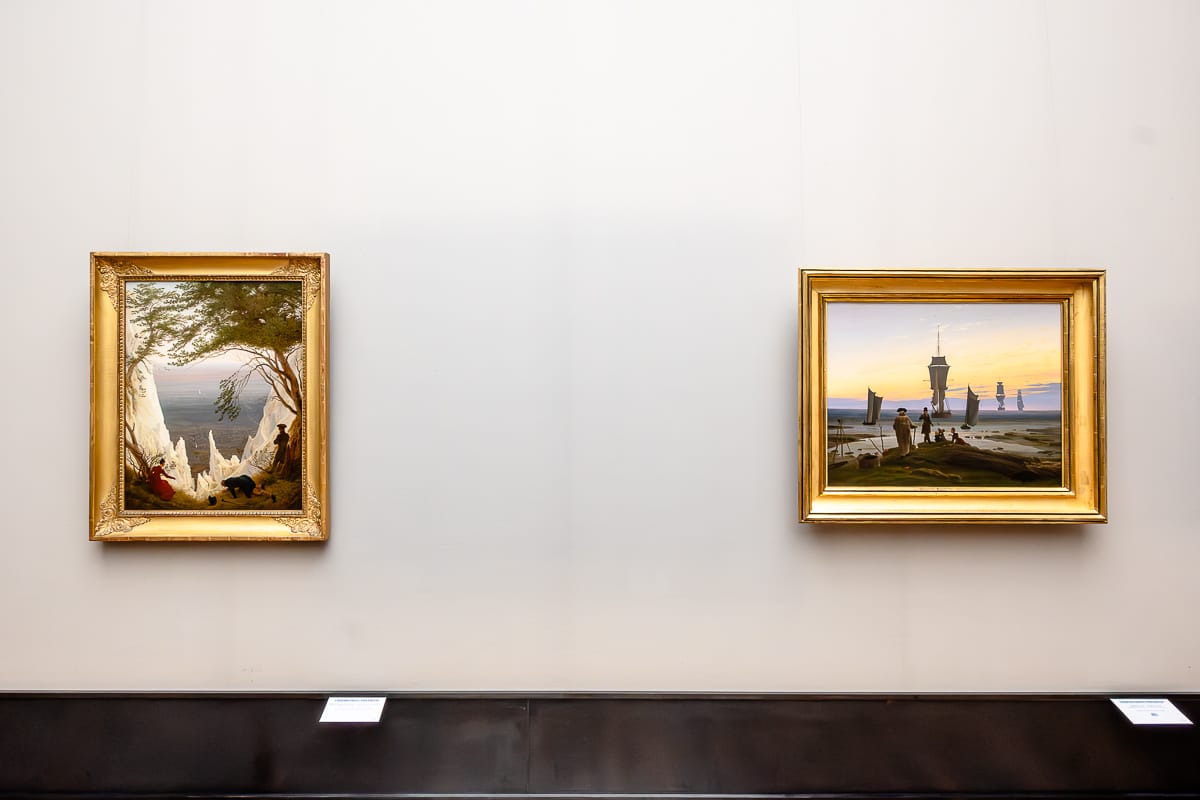 Zwei Bilder hängen etwas weiter auseinander. Links ein größeres Hochformat "Kreidefelsen auf Rügen" (1818) von Caspar David Friedrich, Bäume rahmen das Bild links und rechts, dann Kreidefelsen link sund rechts, drei Personen schauen hinab, eine Frau links zeigt auf etwas runter, ein Mann liegt mittig-rechts am Abgrund und sein Hut links neben ihm, ein weiterer Mann lehnt rechts an einem Baum. Rechts ein größeres Querformat von Caspar David Friedrich, die "Lebensstufen" (um 1834), die am Wasser eine Familie (Mann, Frau, zwei Kinder) und einen älteren Herren, der auf sie zugeht zeigt, im Hintergrund drei große Segler nach hinten versetzt und zwei kleinere Boote, die auch noch auf dem Wasser sind.