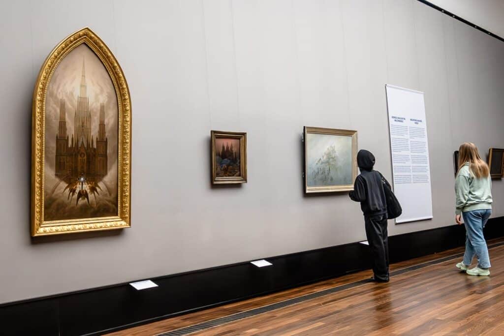 Drei Bilder von Caspar David Friedrich nebeneinander in der Alten Nationalgalerie, ein Informationsplakat dahinter, das Bild davor ("Morgennebel im Gebirge", 1808) wird von einer Frau betrachtet. Mittig kleinformatig und hochkant "Kreuz im Gebirge" (um 1812), ein sehr symmetrisches Bild mit einer Kirche mittig mit zwei Bäumen jeweils rechts und links, davor mittig ein Kreuz, Gebirgssteinformationen. Das linke Bild ist das größte und läuft oben spitz zu, wie ein Altarbild. "Die Kathedrale" (um 1818) mit einer großen Kirche, ein Kreuz davor, Engel darum, ein Regenbogen verläuft über dem Kreuz. 