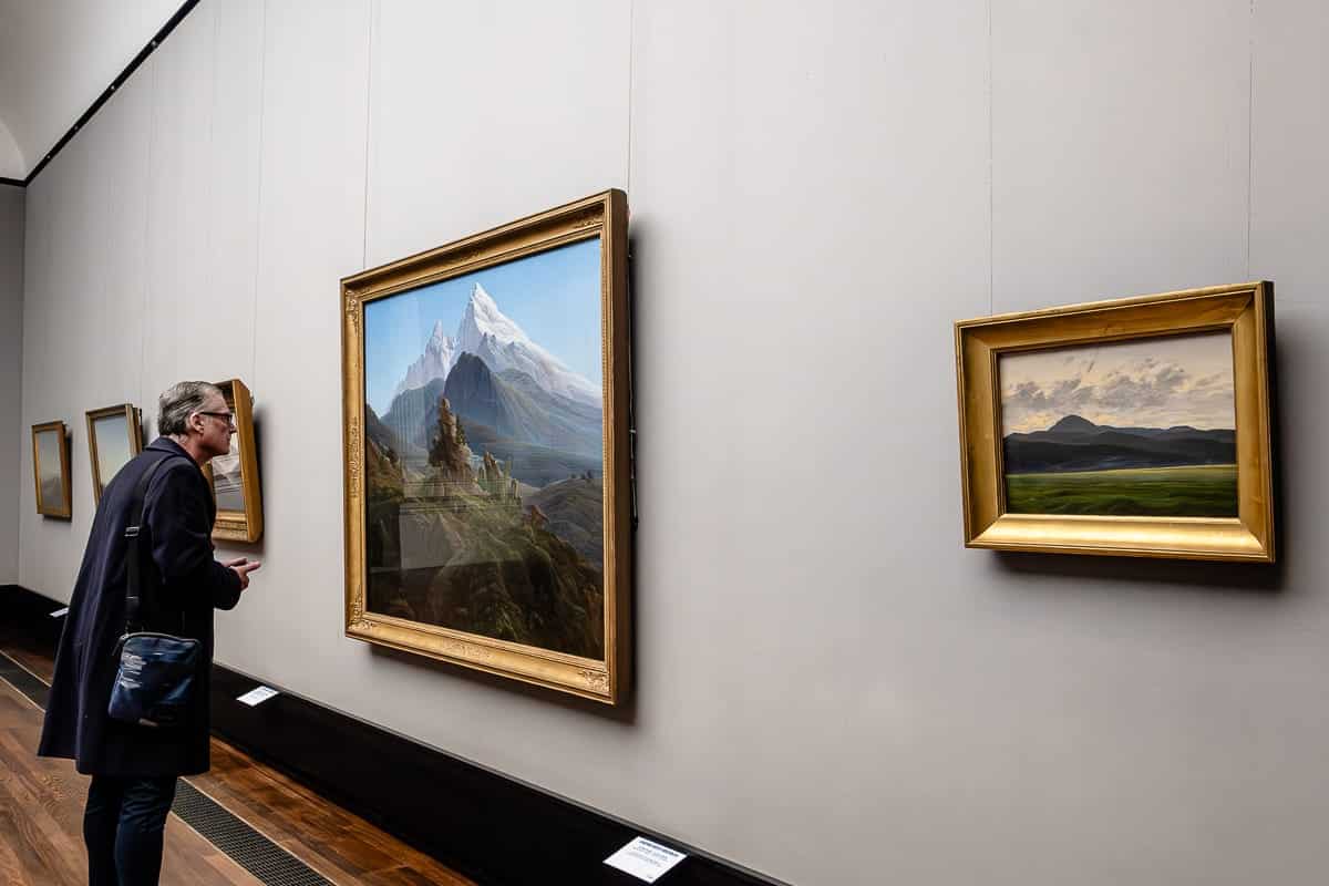 Bilder an einer Wand. Ein sehr großes, querformatiges Bild in der Mitte, ein Mann steht gebeugt davor, um es näher zu betrachten. Auf dem Bild "Der Watzmann" (1824/25) von Caspar David Friedrich in der Alten Nationalgalerie, Berlin ist ein massiver Berg mit weißer Kuppe dargestellt mit den kleineren Bergrücken und Hügeln mit Steinformationen vor ihm.