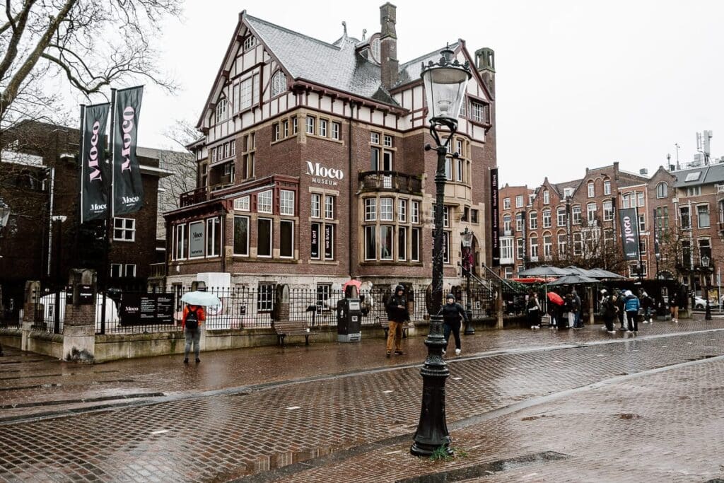 Moco Museum Amsterdam. Seitliche Ansicht von der gegenüberliegenden Seite der Straße, Straßenlaterne im Vordergund, in der linken Ecke des Grundstücks Kunstwerke im Garten, zwei Fahnenmasten mit Moco-Aufdruck, Menschen stehen mit Regenschirmen am Grundstück und am Eingang.