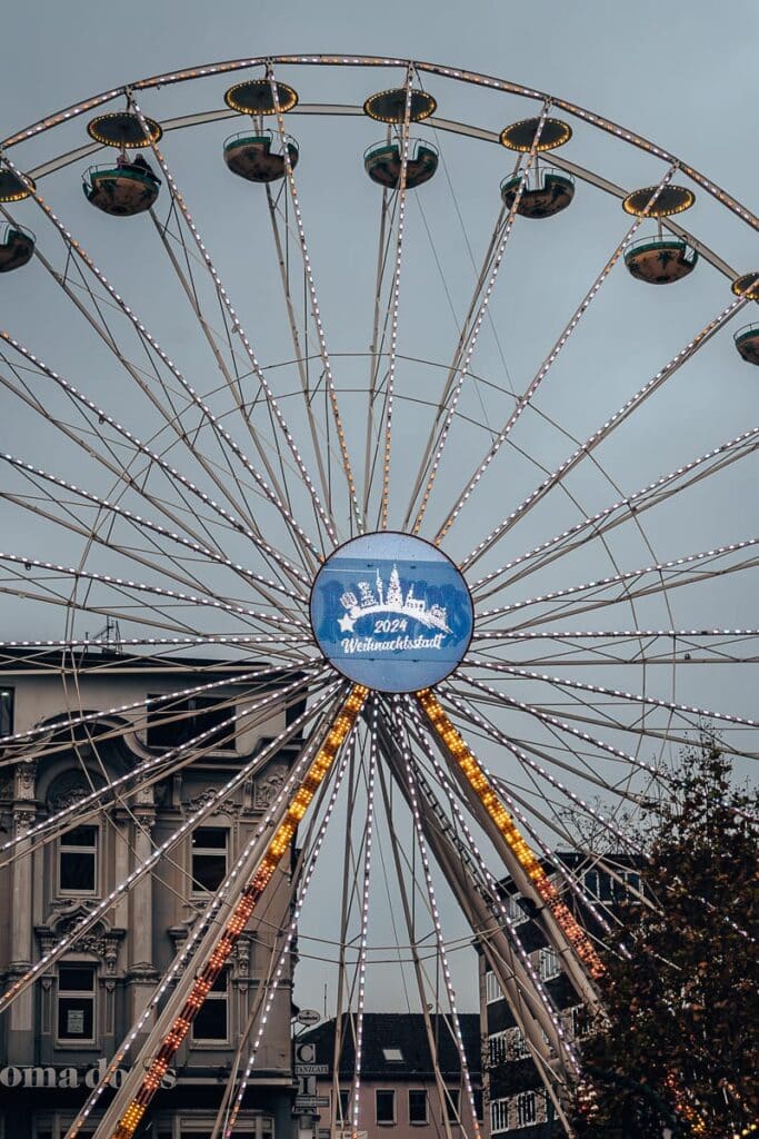 Vor einem bewölkten Himmel steht ein großes, mit Lichtern geschmücktes Riesenrad. In der Mitte ist in weißen Buchstaben ein Schild mit der Aufschrift „2024 Weihnachtsstadt“ angebracht, während in der Nähe das festliche Leuchten eines Weihnachtsbaums der Szenerie Charme verleiht. An der Basis sind die umliegenden Gebäude zu sehen, im Vordergrund das Laub der Bäume.