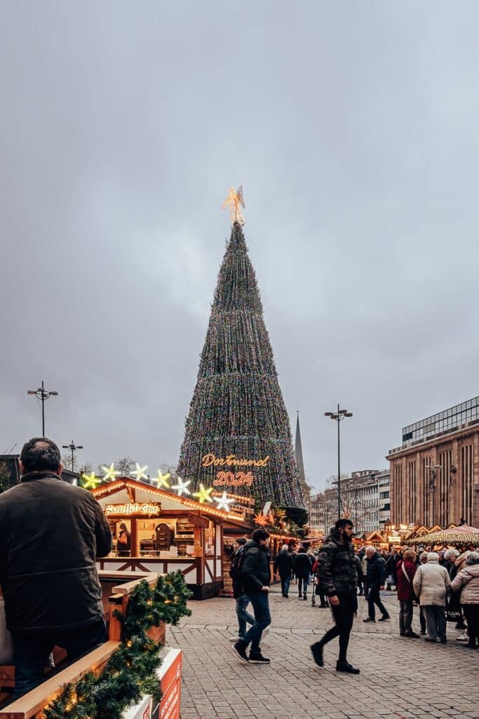 Ein großer Weihnachtsbaum, geschmückt mit Lichtern und einem Engel an der Spitze, steht auf einem geschäftigen Freiluftmarkt in Dortmund. Menschen schlendern umher und besuchen festlich geschmückte Holzstände. Der Himmel ist bedeckt und verbreitet eine gemütliche Winteratmosphäre.