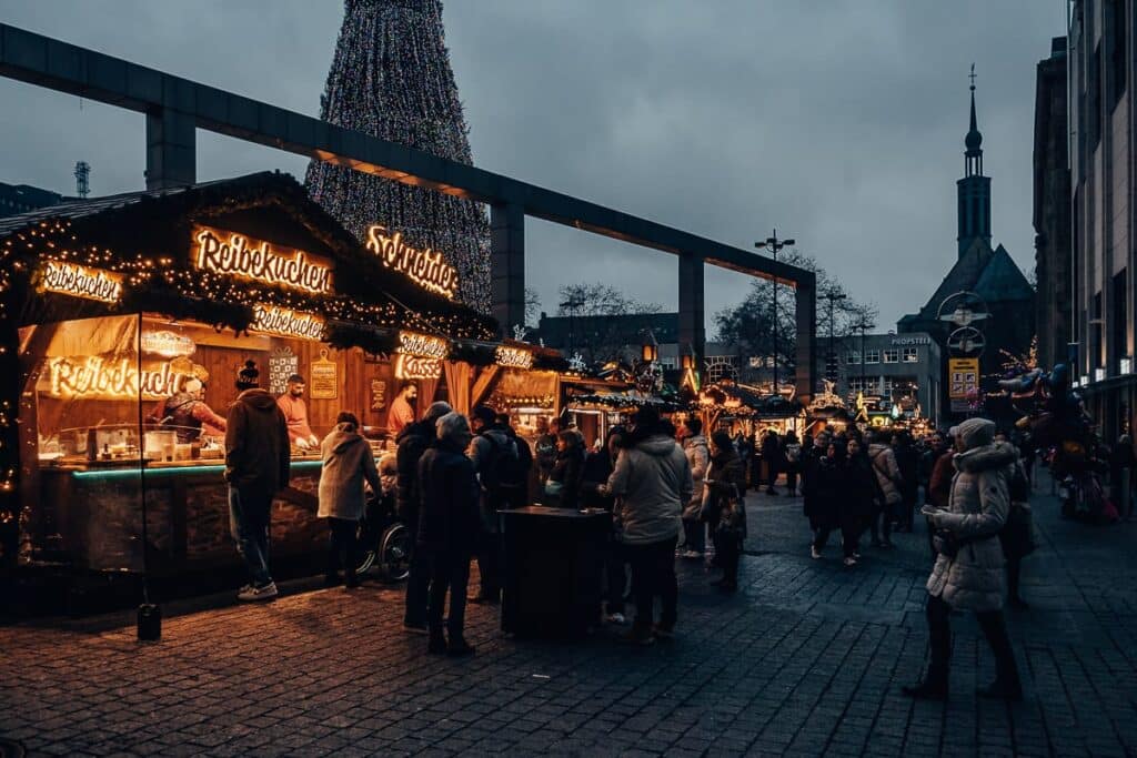 Ein geschäftiger Weihnachtsmarkt im Freien in der Abenddämmerung bietet Essensstände, die mit festlichen Lichtern und Girlanden geschmückt sind. Die Menschen versammeln sich und genießen die lebhafte Atmosphäre. Ein großer Weihnachtsbaum und ein Kirchturm im Hintergrund sorgen für festliche Stimmung vor einem bedeckten Himmel.