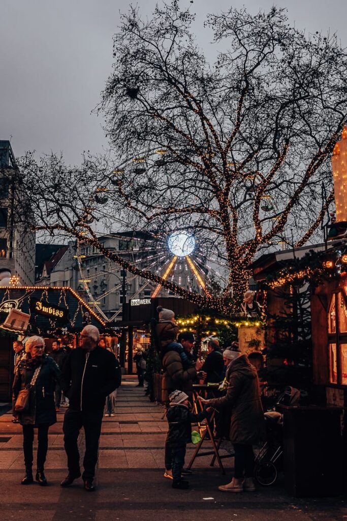 Vor einem bewölkten Himmel steht ein großes, mit Lichtern geschmücktes Riesenrad. In der Mitte ist in weißen Buchstaben ein Schild mit der Aufschrift „2024 Weihnachtsstadt“ angebracht, während in der Nähe das festliche Leuchten eines Weihnachtsbaums der Szenerie Charme verleiht. An der Basis sind die umliegenden Gebäude zu sehen, im Vordergrund das Laub der Bäume.