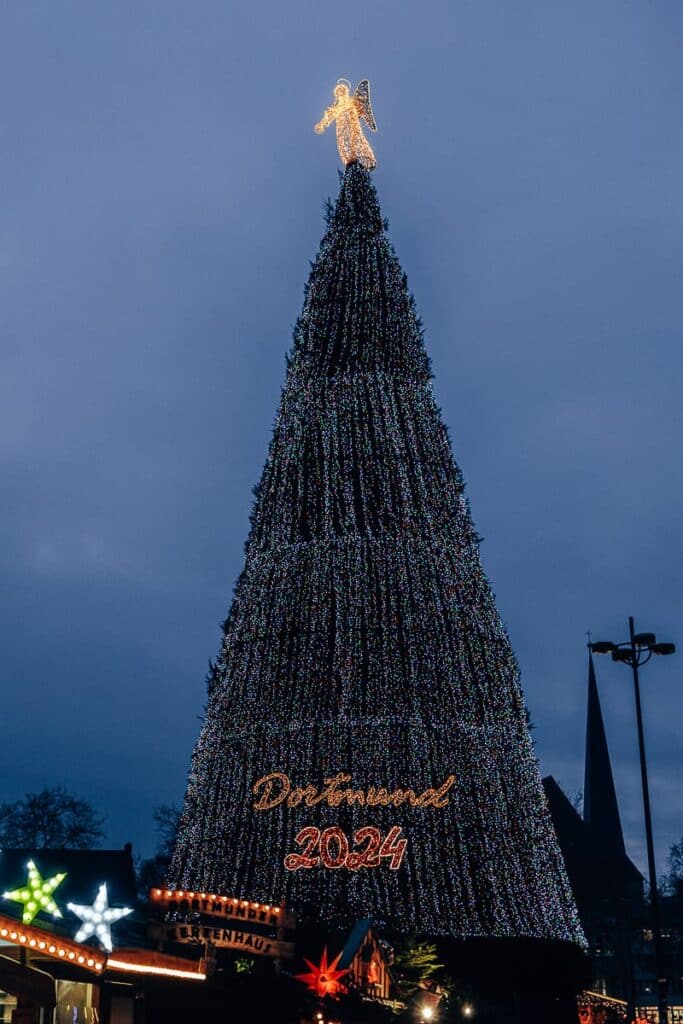 Der hohe Weihnachtsbaum Dortmund, geschmückt mit unzähligen Lichtern und einem leuchtenden Engel als Spitze, dominiert die Szene. An seinem Fuß ist das Schild „Dortmund 2024“ beleuchtet. Der Baum ist von festlichen Ständen mit bunten Lichtern umgeben, die sich vor einem dunklen, bewölkten Abendhimmel abheben.