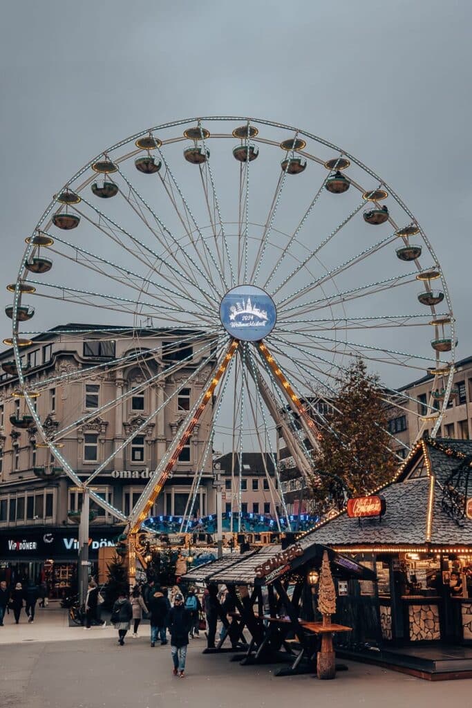 Ein großes Riesenrad ist vor einem bewölkten Himmel in einem Stadtgebiet beleuchtet. Darunter ist ein geschäftiger Weihnachtsmarkt mit dekorierten Ständen mit Lichtern und Girlanden zu sehen. In der Mitte steht ein Weihnachtsbaum, während die Menschen über den Markt schlendern, einkaufen und die festliche Atmosphäre genießen.