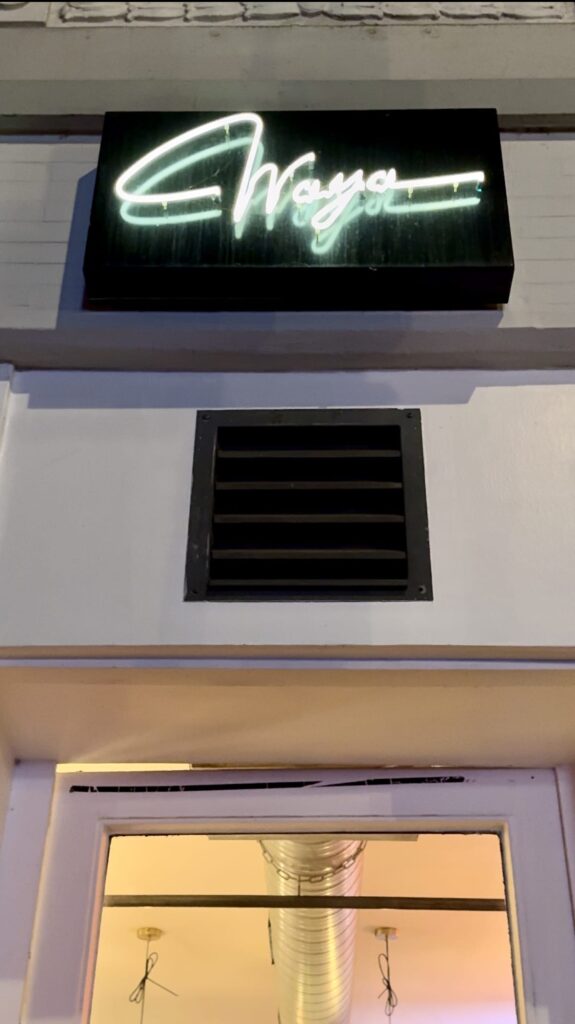 Das Bild zeigt eine Gebäudefassade mit einem Neonschild, auf dem in Schreibschrift „Waya“ steht. Darunter sticht ein schwarzer Lüftungsschacht hervor. Die Glastür zum Eingang bietet einen Blick in das Innere von Waya Kitchen mit sichtbarer Beleuchtung und einem oben angebrachten Lüftungsschacht an der weiß gestrichenen Wand.
