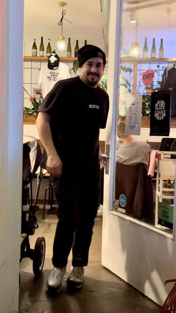 Eine Person mit schwarzer Beanie und „Nester“-T-Shirt lächelt, als sie durch die Tür von Waya Kitchen geht, einem gemütlichen Café, das mit Pflanzen und Flaschen dekoriert ist. Es ist Geschäftsführer und Koch Nick. Drinnen tragen ein weiterer Gast und ein Spaziergänger zur lebhaften Atmosphäre bei. An der Wand fällt ein T-Shirt mit der Aufschrift „Black Coffee“ ins Auge.