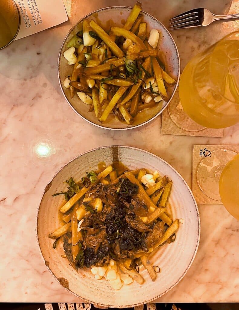 Zwei Teller Poutine von Waya Kitchen stehen auf einem Marmortisch. Der obere Teller enthält Pommes, Käsebruch und Bratensoße, während der untere Teller zusätzliche Beläge wie gehackte Kräuter und karamellisierte Zwiebeln enthält. Eine Gabel und ein gelbes Getränk liegen neben den Tellern auf beigen Untersetzern.