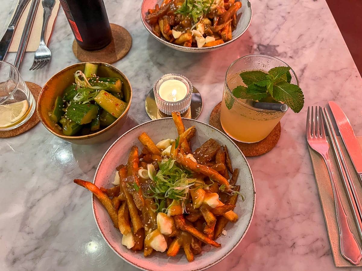 Eine Draufsicht auf einen Waya Kitchen-Tisch zeigt zwei Schüsseln Poutine mit gehackten Frühlingszwiebeln, dazu marinierten Gurkenscheiben und einem mit Minze garnierten Cocktail. Eine brennende Kerze und Untersetzer ergänzen die graue Marmoroberfläche, komplett mit elegantem Besteck.