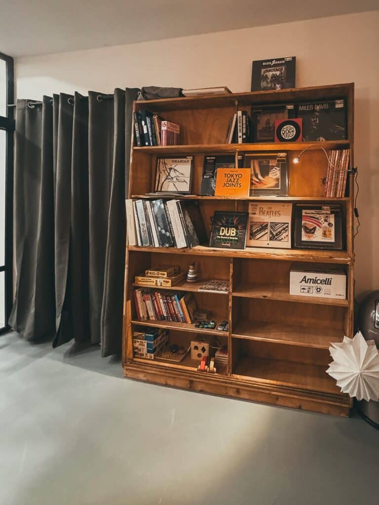 Ein hölzernes Bücherregal an einer Wand ist mit Schallplatten, Büchern und Dekoartikeln gefüllt und verkörpert die Atmosphäre des mono Listening Cafés, in dem man Musik hören kann. Einige Buchtitel sind im sanften Schein einer kleinen Lampe auf einem unteren Regal zu sehen. Ein großes Fenster mit grauen Vorhängen verstärkt die minimalistische und gemütliche Atmosphäre.