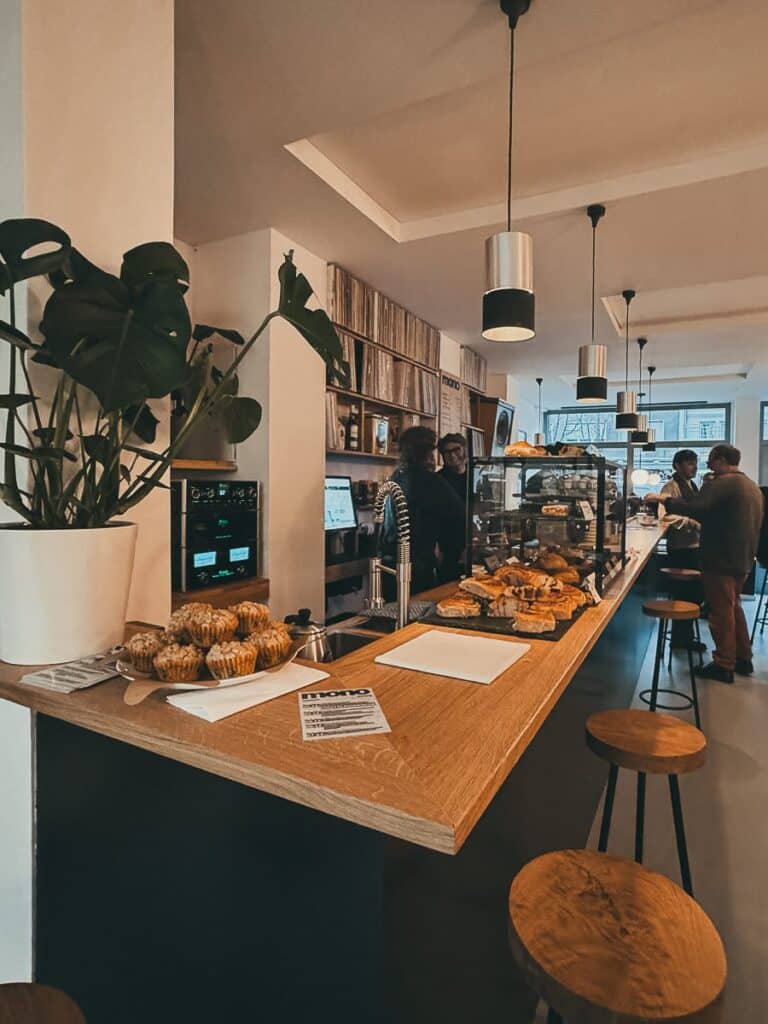In einem gemütlichen Café ist die Holztheke mit Muffins und Gebäck geschmückt. Barhocker laden die Gäste zum Entspannen ein, während eine Topfpflanze für Grün sorgt. Im Hintergrund unterhalten sich die Leute neben einer Gebäckvitrine unter warmen Hängelampen und schaffen so eine einladende Atmosphäre, die perfekt zum ruhigen Zuhören ist.