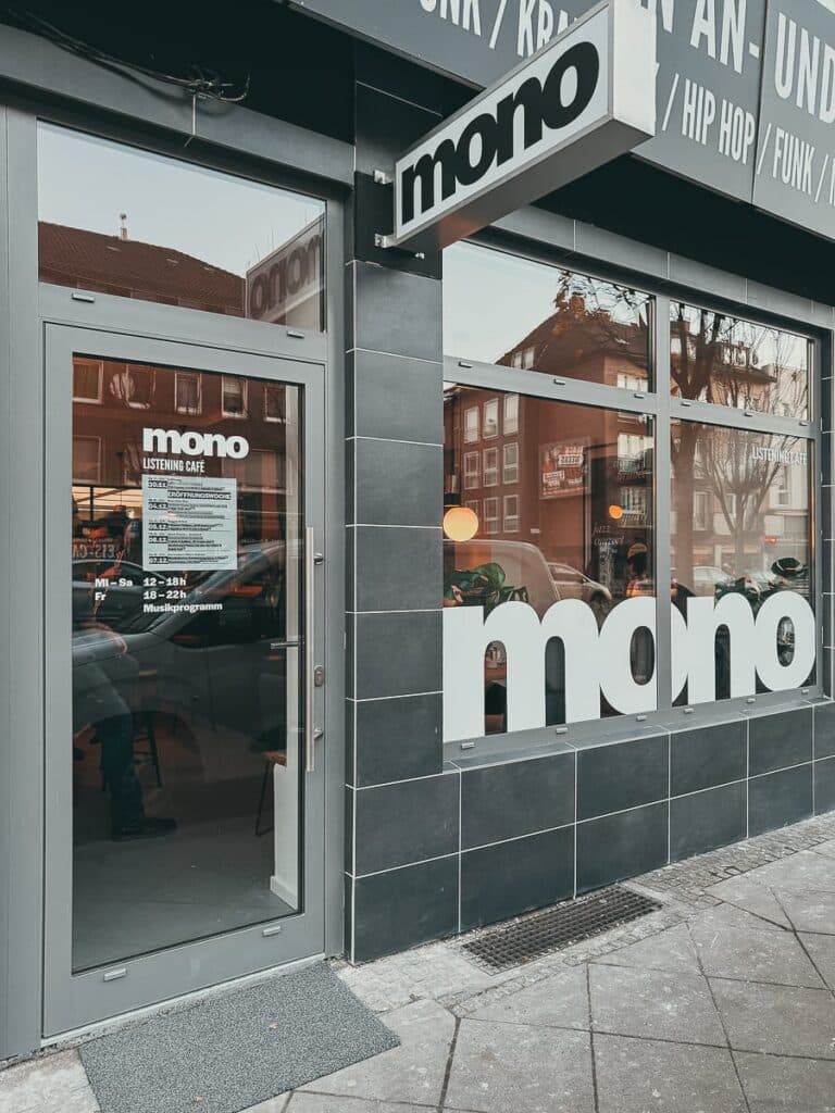 Eine moderne Ladenfront mit dunklen Fliesen, großen Fenstern und „mono“ Beschilderung in Weiß. Dieses einladende Café zum Zuhören verfügt über ein Türschild mit den Öffnungszeiten: Montag bis Sonntag, 10 bis 18 Uhr, mit einem Freitagsmusikangebot von 18 bis 22 Uhr. Im Glas spiegeln sich die umliegenden Gebäude und Passanten.