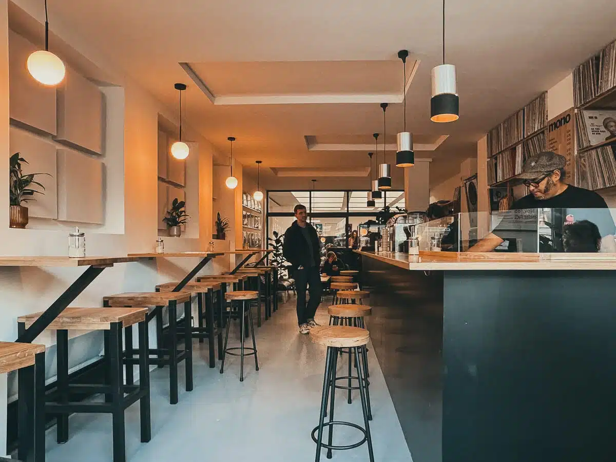 Ein gemütliches Café-Interieur mit Holztischen und Hockern auf beiden Seiten. Warme Hängelampen sorgen für Atmosphäre. Eine Person geht den Gang entlang, eine andere steht rechts hinter der schwarzen Theke. Die Wände sind mit Pflanzen und Regalen voller Schallplatten dekoriert, die für einen monochromen Touch sorgen, während große Fenster auf der Rückseite natürliches Licht hereinlassen.