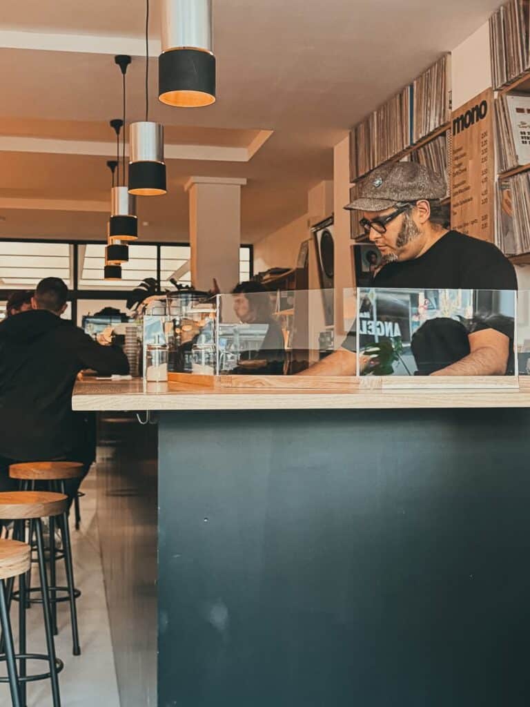 Ein DJ mit Mütze und Sonnenbrille legt hinter der Theke eines Cafés mit minimalistischem Dekor auf. Die hölzerne Theke mit hohen Hockern verleiht dem Ganzen Charme, während Deckenleuchten die sich unterhaltenden Gäste sanft beleuchten.