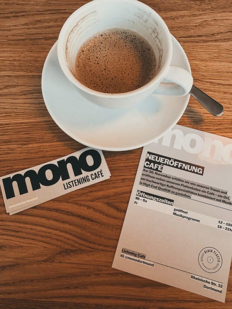 Auf einem Holztisch steht eine weiße Kaffeetasse mit Espresso auf einer Untertasse. Daneben fallen eine Visitenkarte und ein Flyer ins Auge. Der Flyer kündigt die Eröffnung des MONO Listening Café an, nennt den Standort in der Rheinischen Straße 32 und nennt die Öffnungszeiten.