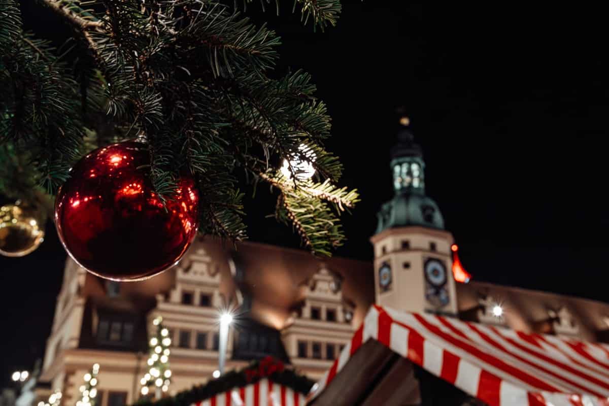 Eine festliche Szene mit einer Nahaufnahme eines roten Weihnachtsornaments und Lichtern, die von einem Tannenzweig hängen. Im Hintergrund ist ein historisches Gebäude mit einem Uhrturm nachts beleuchtet. Darunter lassen gestreifte rot-weiße Dächer eine Weihnachtsmarktkulisse vermuten.