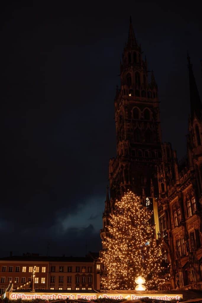 Ein großer, hell erleuchteter Weihnachtsbaum steht nachts vor einem reich verzierten, dunklen Gebäude im gotischen Stil - Rathaus München. Der Himmel ist bewölkt und festliche Lichter erhellen die Szene und schaffen eine warme, einladende Atmosphäre. Am Fuß des Baumes erscheinen aus einer Lichterkette deutsche Sätze.