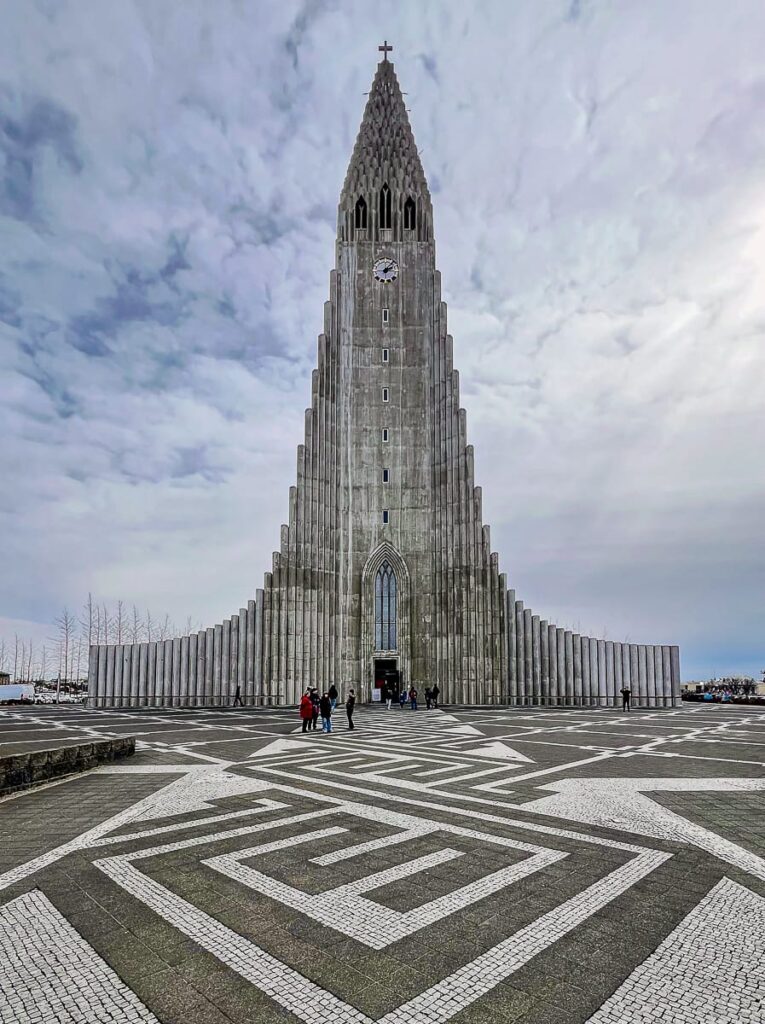 Eine hohe, moderne Betonkirche mit abgestufter Fassade und spitzem Turm (Hallgrímskirkja) ragt in einen wolkigen Himmel von Reykjavík. Der Eingang ist von Säulen flankiert. Im Vordergrund ziert ein geometrisches Muster den Platz, auf dem ein paar Menschen spazieren gehen. Vereinzelte Bäume säumen den linken Hintergrund.