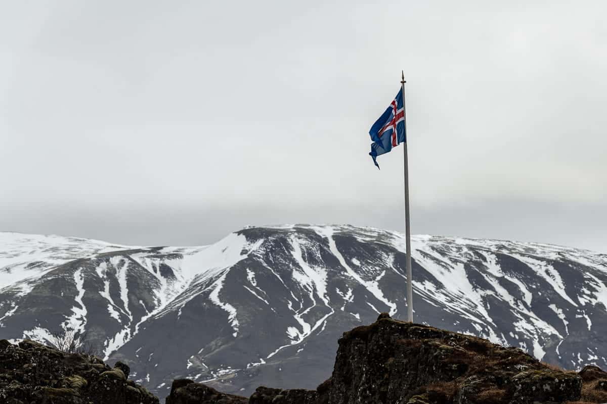 Eine Flagge an einem Mast weht im Wind, in den Farben Blau, Rot und Weiß der isländischen Nationalflagge. Sie ist vor einer Kulisse aus schneebedeckten Bergen und einem bewölkten Himmel zu sehen, wodurch ein starker Kontrast zwischen der rauen Landschaft und den leuchtenden Farben der Flagge entsteht.