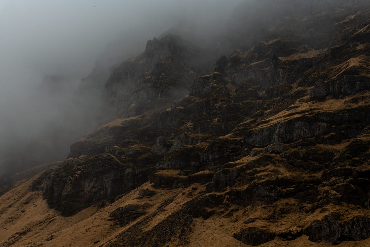 Nebliger Berghang mit dunklem, zerklüftetem Gelände, das teilweise mit braunem Gras bedeckt ist auf Island. Nebel hüllt die felsigen Klippen ein und schafft eine geheimnisvolle und stimmungsvolle Atmosphäre. Der Kontrast zwischen dem Nebel und den dunklen Felsen unterstreicht die Steilheit und Beschaffenheit der Landschaft.