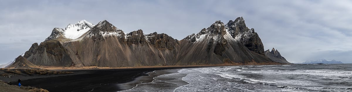 Panoramablick auf den Berg Vestrahorn in Island mit schroffen, teilweise schneebedeckten Gipfeln. Unten erstreckt sich ein schwarzer Sandstrand entlang der Küste, an dessen Ufer sanfte Wellen plätschern. Der Himmel ist bedeckt und wirft ein sanftes, gedämpftes Licht auf die Landschaft.