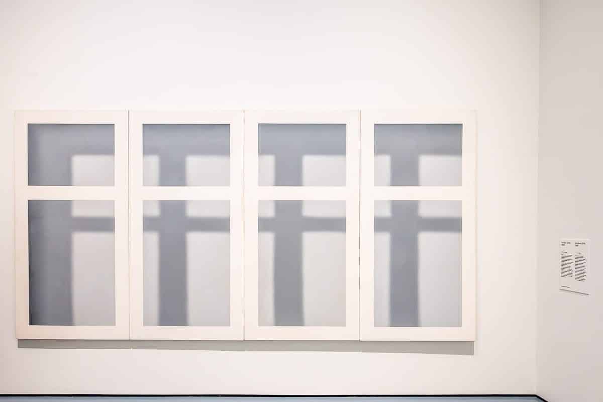 Ein minimalistisches Kunstwerk von Gerhard Richter mit vier vertikalen rechteckigen Tafeln, von denen jede mit drei vertikalen und drei horizontalen Linien gitterartige Strukturen bildet und werfen weiche Schatten. Rechts neben dem Kunstwerk ist ein kleines Etikett mit Text zu sehen.