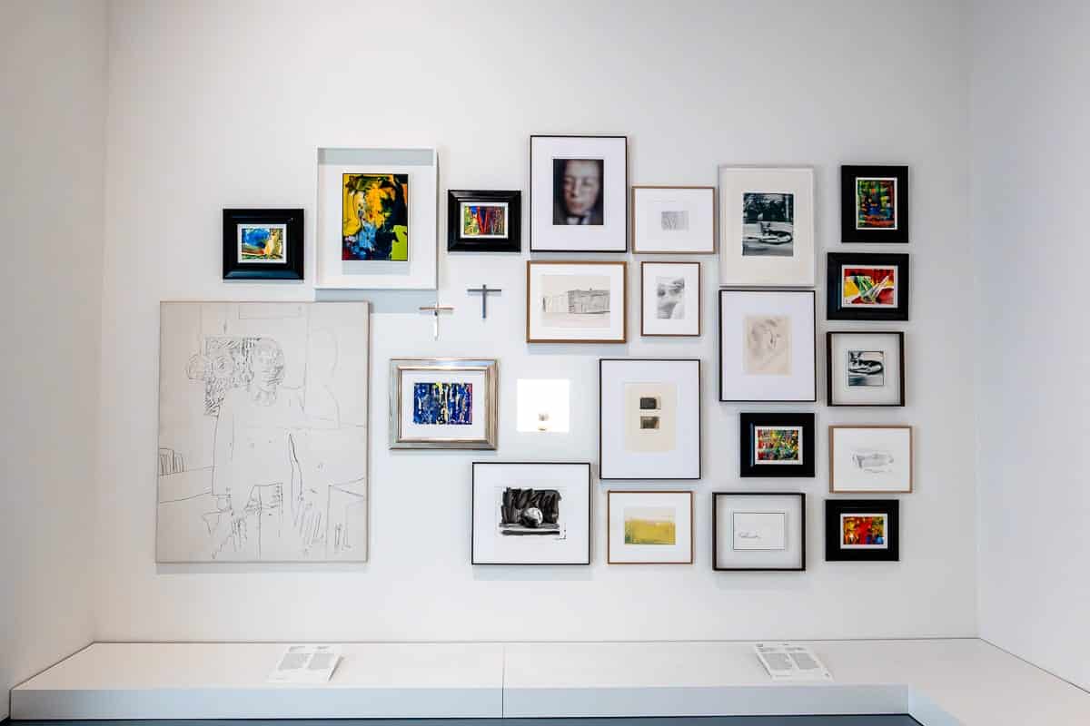 Eine Galeriewand mit einer vielfältigen Anordnung gerahmter Kunstwerke von Gerhard Richter, darunter Skizzen, abstrakte Gemälde, Fotografien und Farbdrucke. Die Rahmen variieren in Größe und Farbe und ergeben eine vielseitige Präsentation. Auf einer kleinen Leiste darunter befinden sich zwei offene Bücher mit sichtbarem Text und Bildern.