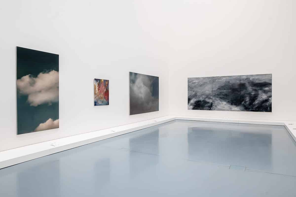 Eine minimalistische Kunstgalerie verfügt über einen reflektierenden Boden, der an ruhiges Wasser erinnert. Vier Gemälde von Gerhard Richter mit abstrakten Wolken- und Himmelsmotiven sind auf weißen Wänden ausgestellt. Die ruhige Atmosphäre wird durch eine gedämpfte Farbpalette aus Blau-, Grau- und sanften Weißtönen verstärkt.