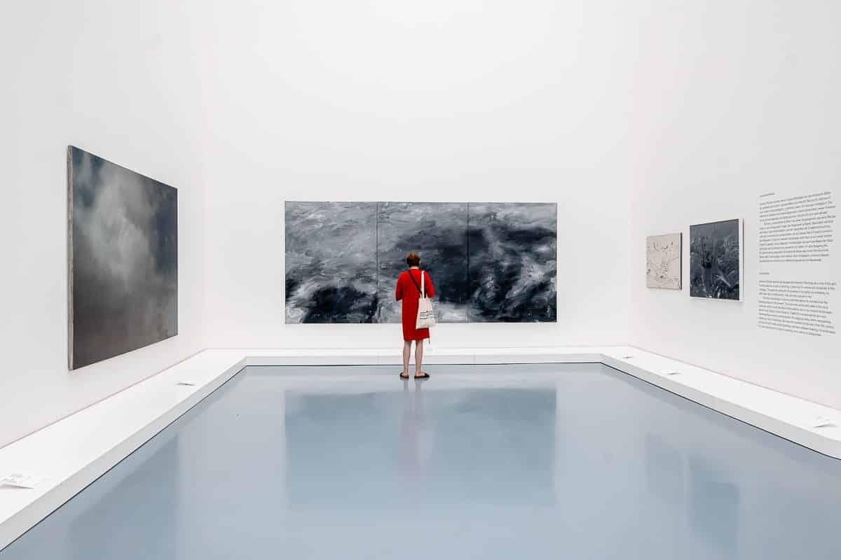 Eine Person in einem roten Outfit steht im Ausstellungsraum des Kunstpalastes Düsseldorf und betrachtet ein großes abstraktes Gemälde von Gerhard Richter mit dunklen, wirbelnden Mustern. Der Boden ist hellblau und die Wände sind weiß und mit mehreren anderen Kunstwerken und einem Textblock bedeckt. Die Person trägt eine weiße Einkaufstasche.