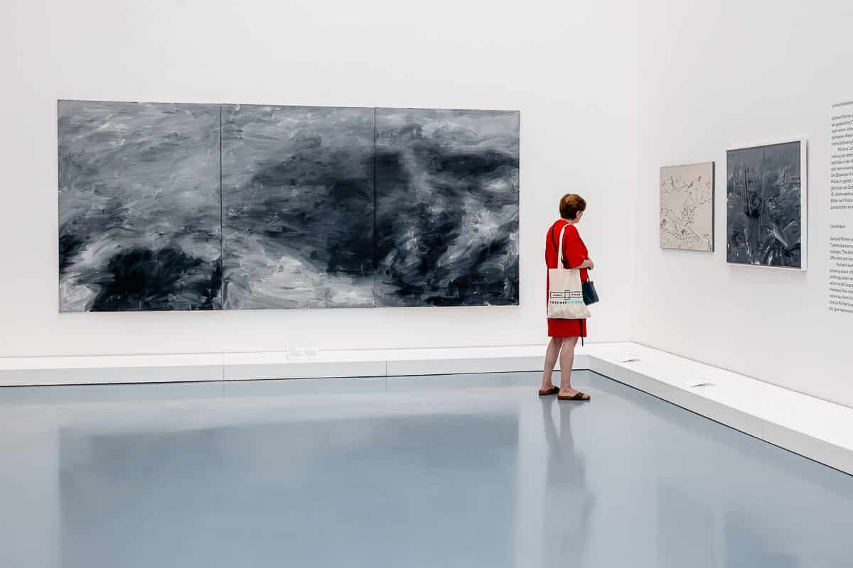 Eine Person in einem roten Kleid und Sandalen steht in einer Kunstgalerie und betrachtet ein großes abstraktes Gemälde von Gerhard Richter in Graustufen und kleinere gerahmte Werke. Sie trägt eine Einkaufstasche und eine blaue Tasche. Der Raum hat einen polierten hellgrauen Boden und strahlend weiße Wände. An der Wand ist Text zu sehen.
