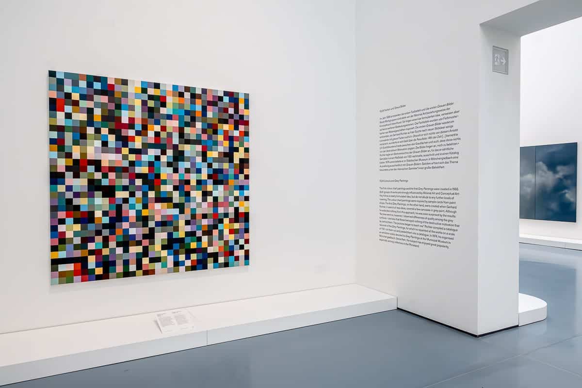 Eine Galerie für moderne Kunst zeigt ein farbenfrohes abstraktes Gemälde aus kleinen, bunten Quadraten auf einer weißen Wand von Gerhard Richter. Rechts auf einer weißen Wand befindet sich ein beschreibender Text zum Kunstwerk. Ein weiteres verschwommenes Gemälde ist teilweise durch eine offene Tür sichtbar, alles auf einem glänzenden grauen Boden.
