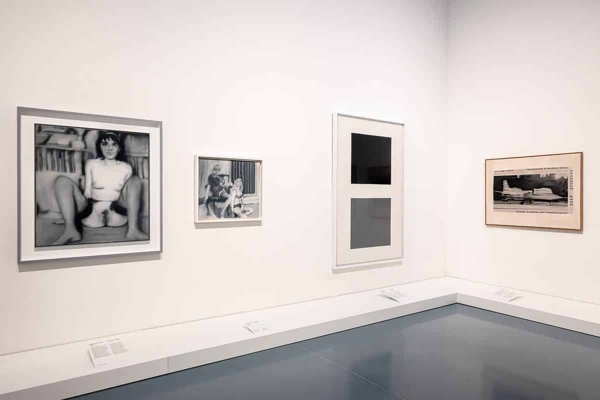 Ein Galerieraum mit vier gerahmten Kunstwerken von Gerhard Richter an weißen Wänden. Die ersten beiden links sind Schwarzweißfotos von Menschen. Das dritte ist ein modernes abstraktes Werk mit Rechtecken. Das vierte ist ein Sepiabild eines Flugzeugs. Der Boden ist glänzend grau und unter jedem Kunstwerk befinden sich Etiketten.