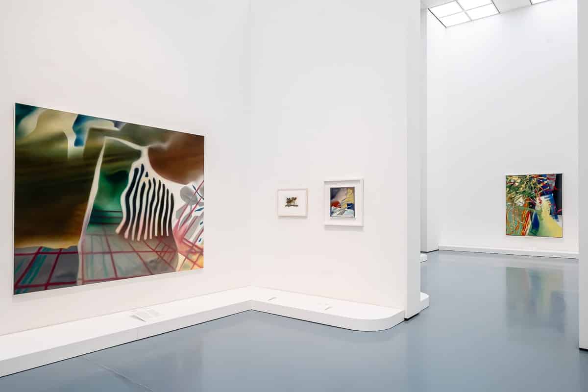 Ausstellung "Verborgene Schätze", Werke von Gerhard Richter aus Privatsammlungen im Rheinland, im Kunstpalast Düsseldorf mit einem großen abstrakten Gemälde an einer Wand mit Wellenlinien und leuchtenden Farben. Daneben befinden sich zwei kleinere gerahmte Kunstwerke. Im Hintergrund ist ein weiteres großes abstraktes Gemälde zu sehen. Der Raum ist minimalistisch gestaltet, mit hohen Decken und polierten grauen Böden.