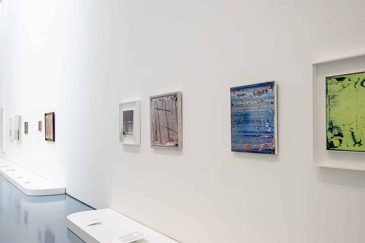 Ausstellungsraum des Kunstpalasts Düsseldorf mit hohen weißen Wänden, in der abstrakte Gemälde von Gerhard Richter in verschiedenen Farben und Größen ausgestellt sind. Die Gemälde sind gerahmt und in gleichmäßigen Abständen angeordnet. Der Boden ist in glänzendem Grau gehalten und neben den Kunstwerken befinden sich Beschreibungen. Die Perspektive führt den Blick weiter den Korridor hinunter.