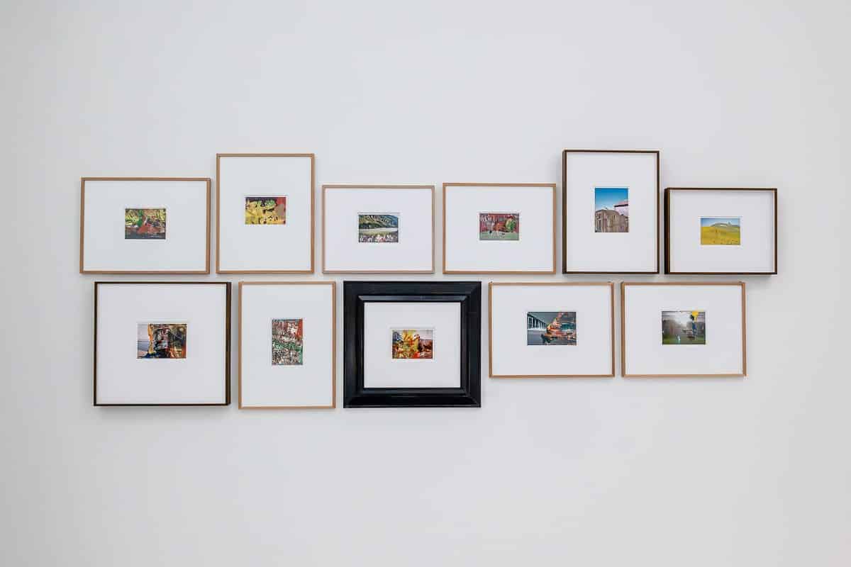 Eine Sammlung von 11 gerahmten Fotos von Gerhard Richter, die an einer weißen Wand angeordnet sind. Die meisten Rahmen sind aus Holz mit weißen Matten, mit Ausnahme eines einzelnen schwarzen Rahmens in der Mitte. Die Bilder in den Rahmen zeigen verschiedene Szenen, darunter Landschaften und abstrakte Kunst. Die Rahmen bilden ein gitterartiges Muster.