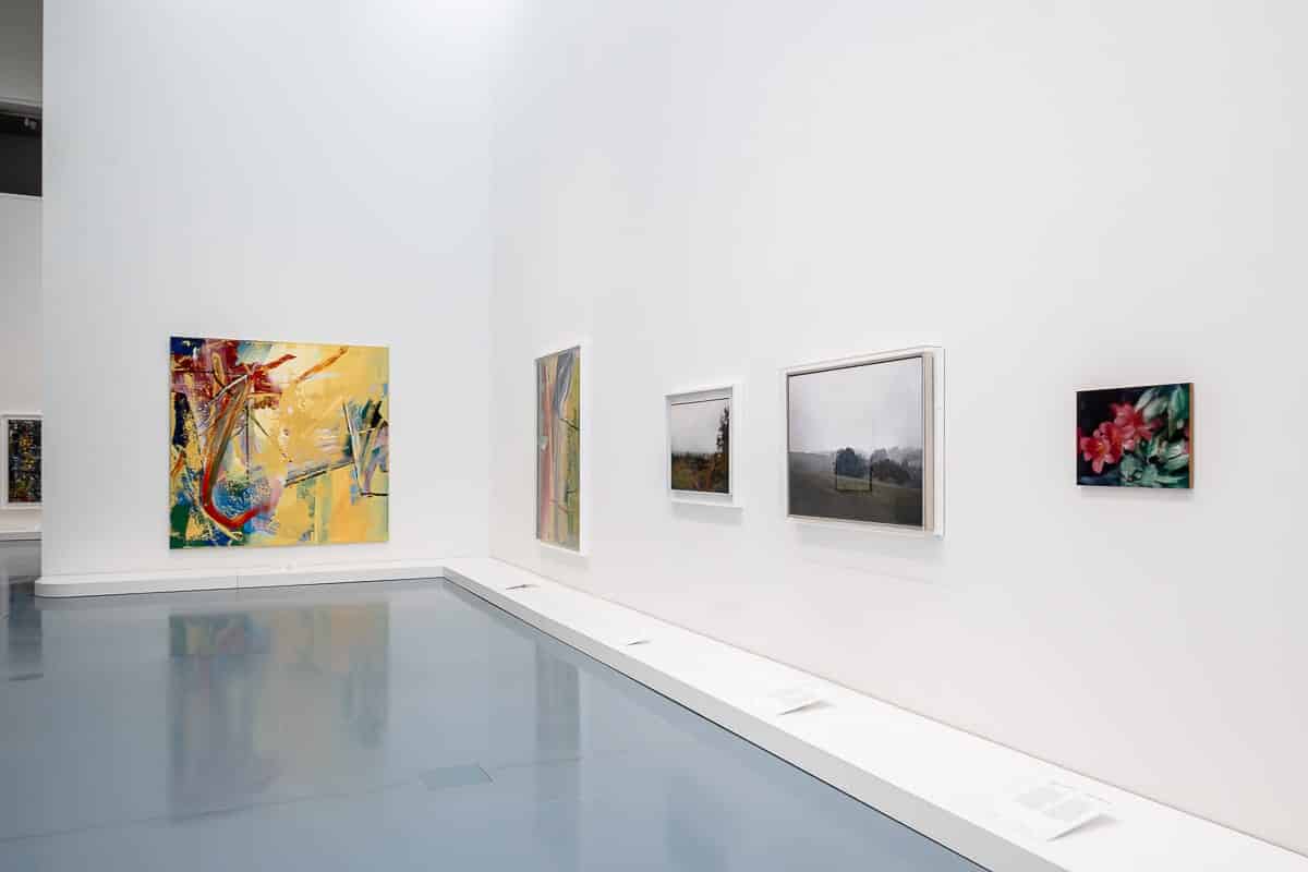 Der Kunstpalast Düsseldorf zeigt sechs Gemälde von Gerhard Richter an einer weißen Wand mit einem glänzenden grauen Boden. Die Kunstwerke variieren in Stil und Größe und zeigen abstrakte und Landschaftsmotive. Das größte, links, ist farbenfroh und abstrakt. Unter jedem Gemälde befinden sich Informationstafeln.
