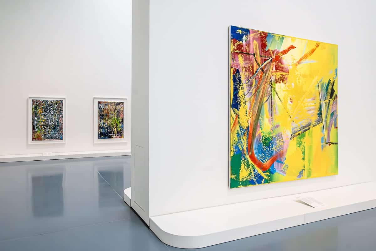 Kunspalast Düsseldorf mit lebendigen abstrakten Gemälden. Ein großes, farbenfrohes Kunstwerk mit dynamischen gelben, blauen und roten Pinselstrichen ist prominent an einer weißen Wand ausgestellt. Im Hintergrund hängen zwei gerahmte Kunstwerke mit komplizierten Mustern an einer weißen Wand. Der Boden ist glänzend grau.