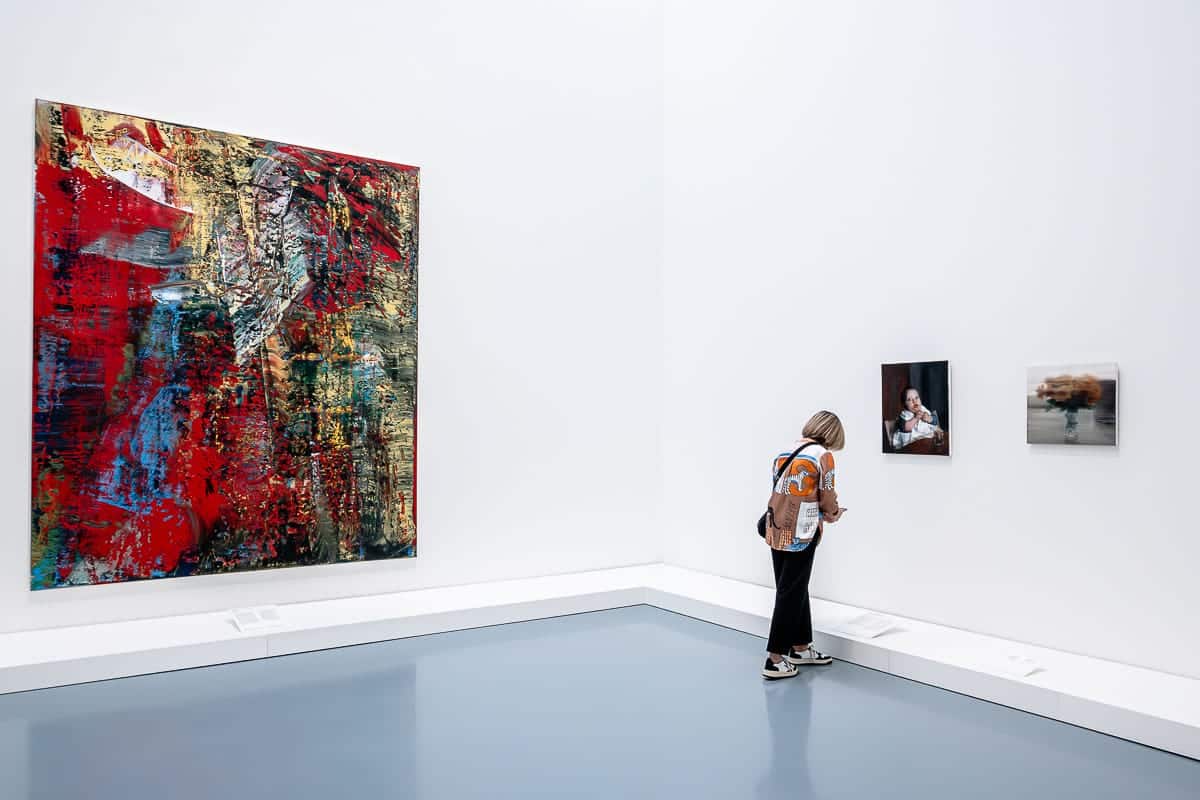 Eine Person steht im letzten Ausstellungsraum im Kunstpalas Düsseldorf der Ausstellung Gerhard Richter "Verborgene Schätze" mit weißen Wänden und blaugrauem Boden und betrachtet ein Kunstwerk. Links hängt ein großes abstraktes Gemälde in den Farben Rot, Blau und Beige. Rechts hängen zwei kleinere Gemälde. Der Betrachter trägt einen Rucksack und eine schwarze Hose.