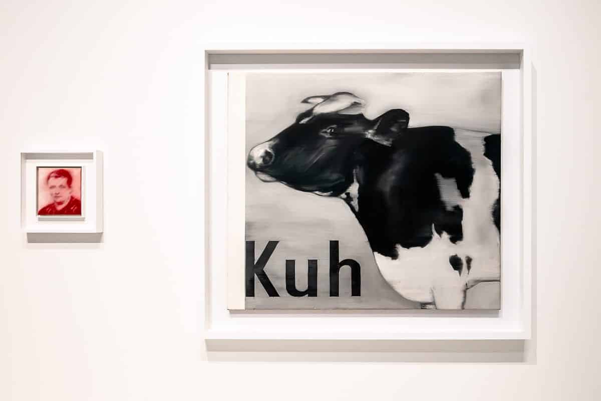 An einer weißen Wand hängt ein Gemälde, auf dem eine schwarz-weiße Kuh mit dem Wort „Kuh“ in fetten Buchstaben abgebildet ist. Daneben hängt ein kleineres, gerahmtes, rot getöntes Porträt einer Person. Beide Stücke sind weiß gerahmt, was ein minimalistisches Bild ergibt. Beide Bilder sind Frühwerke von Gerhard Richter, ausgestellt im Kunstpalast Düsseldorf.