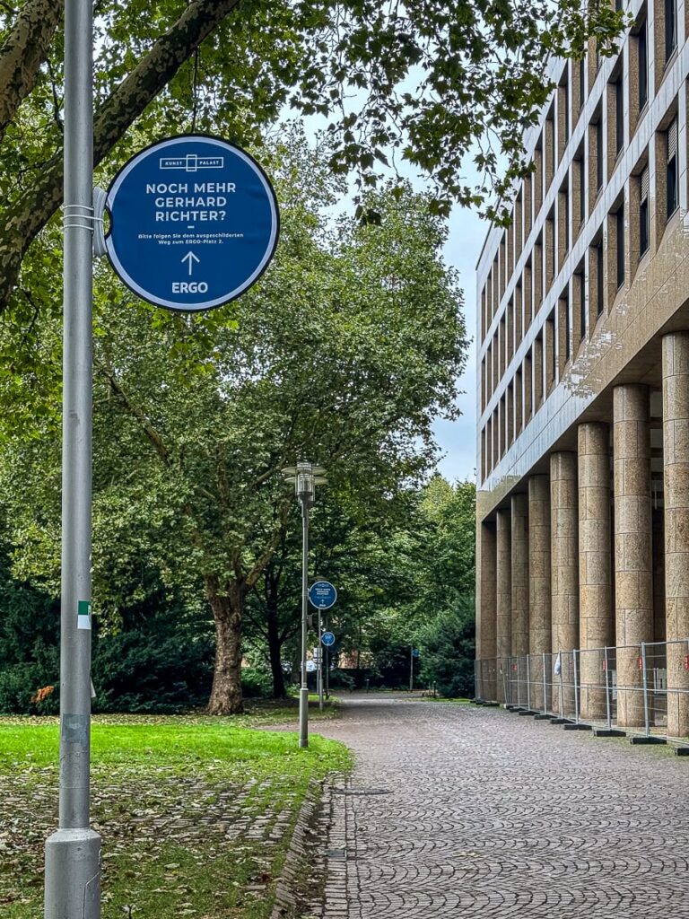 Weg neben einem modernen Gebäude mit Steinsäulen. Bäume mit grünen Blättern säumen den Weg. Auf einem blauen runden Schild steht „Noch Mehr Gerhard Richter?“ mit einem nach vorne zeigenden Pfeil. Der Himmel ist bedeckt und die Umgebung wirkt ruhig und grün.