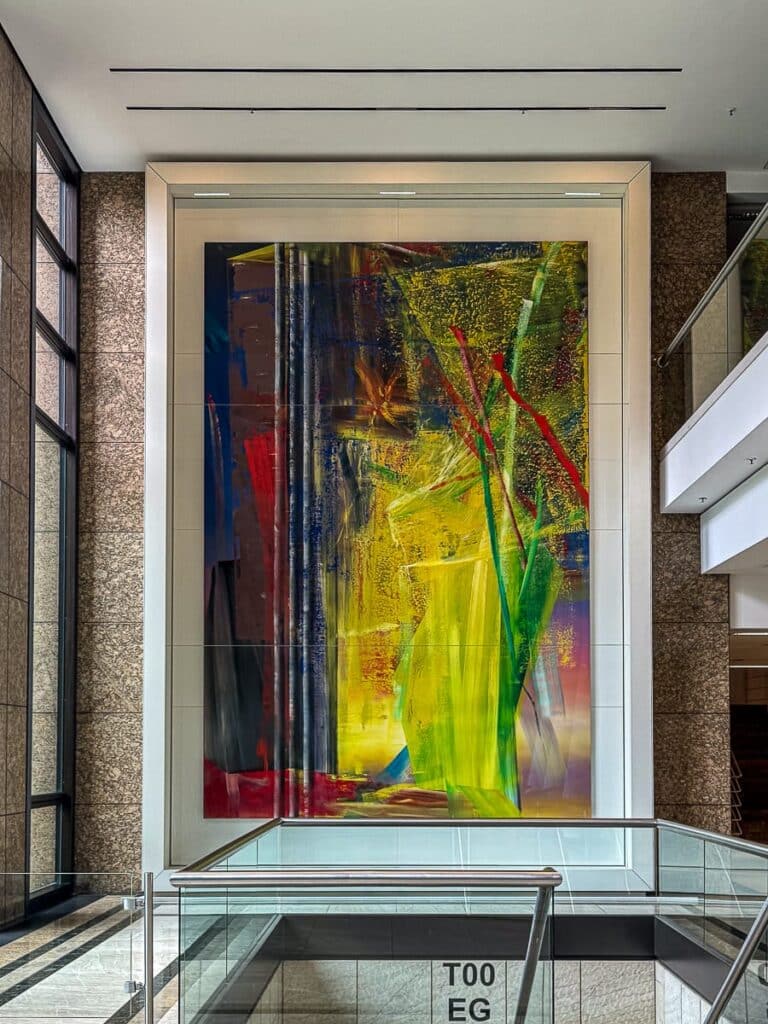 Ein großes abstraktes Gemälde von Gerhard Richter im Foyer eines modernen Gebäudes zeichnet sich durch leuchtende Farben aus, vorwiegend Gelb, Rot und Blau, mit dynamischen Pinselstrichen und Texturen. Es wird von einer hohen, weißen Struktur eingerahmt und über einem Glasgeländer zwischen Steinwänden angebracht.