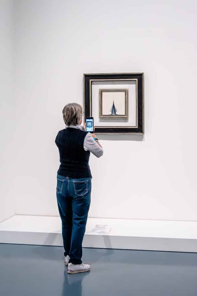 Eine Person mit kurzen Haaren, einer schwarzen Weste, einem weißen Hemd und blauen Jeans steht vor einem gerahmten Gemälde in einer Galerie. Sie hält ein Smartphone hoch, um ein Foto zu machen. Das Gemälde von Gerhard Richter zeigt ein abstraktes, minimalistisches Design mit einer großen, dunklen Form auf einem hellen Hintergrund.