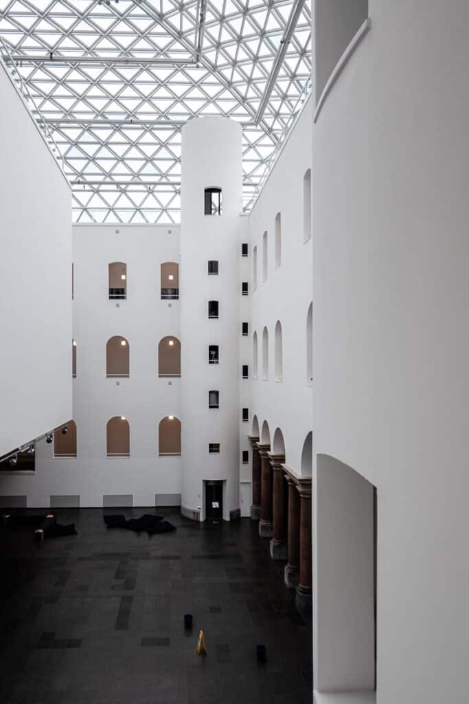 Das geräumige Atrium des K21. Es verfügt über eine Glasdecke und weiße Wände, die durch einen schwarz gefliesten Boden kontrastiert werden. Bogenfenster säumen jede Ebene, während rechts neben dunklen Säulen eine hohe zylindrische Struktur mit winzigen Fenstern die Aufmerksamkeit auf sich zieht. Auf dem Boden steht unauffällig ein gelbes Warnschild. 