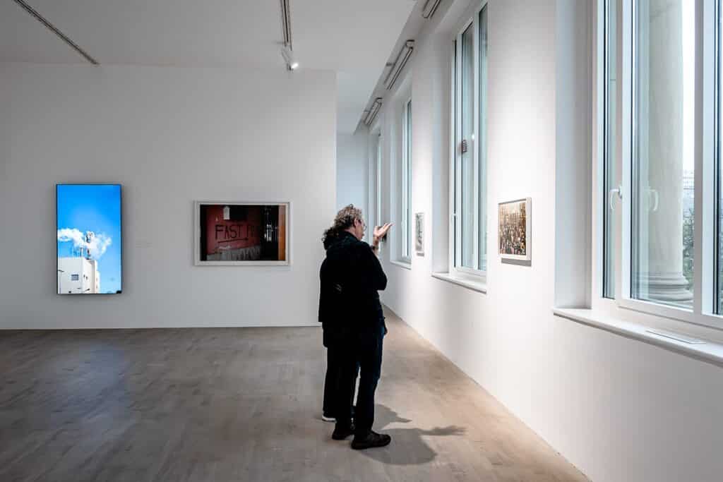 Zwei Menschen stehen eng beieinander in einem hell erleuchteten Raum im K21 Düsseldorf und bewundern gerahmte Fotografien von Lars Eidinger an den weißen Wänden. Große Fenster auf der rechten Seite lassen natürliches Licht herein. 