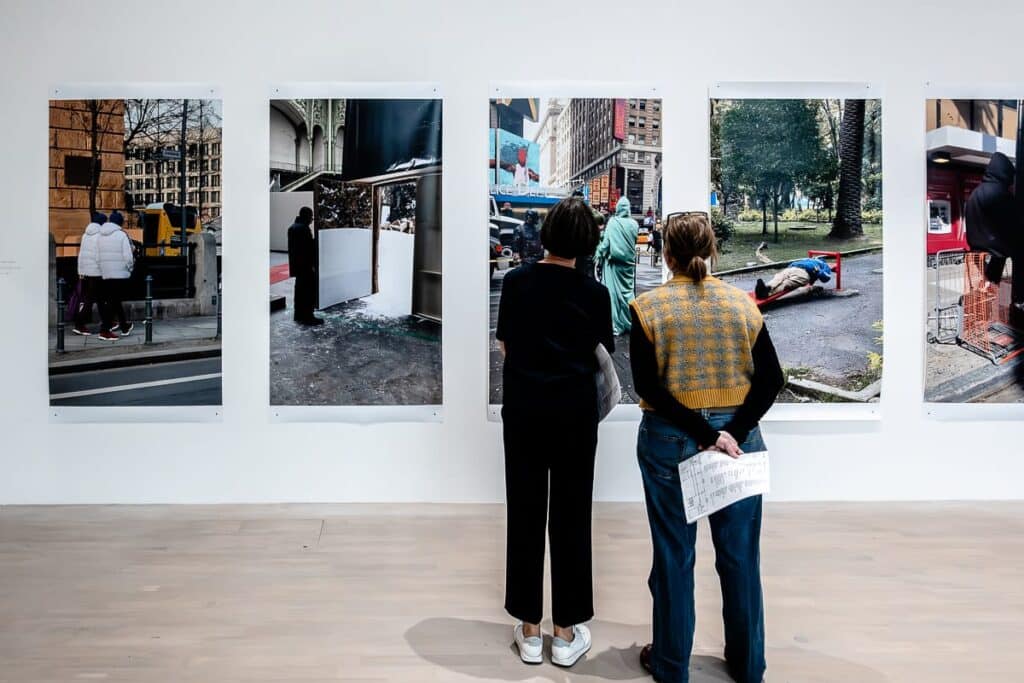 Zwei Personen stehen in einer Galerie und betrachten fünf große, vertikale Fotografien an einer weißen Wand. Die Bilder zeigen verschiedene urbane Szenen und Gebäude. Ein Betrachter hält ein Blatt Papier in der Hand, beide sind mit den Kunstwerken beschäftigt.