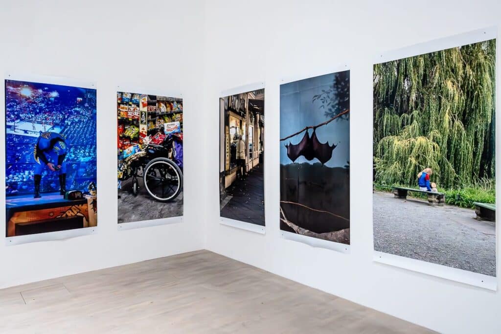 Ein Galerieraum mit fünf großen Fotografien auf weißen Wänden. Jedes Foto zeigt eine Person, die sich von der Kamera abwendet und in alltägliche Aktivitäten vertieft ist, vor unterschiedlichen städtischen und natürlichen Hintergründen. Die Umgebung ist minimalistisch, mit hellem Holzboden.