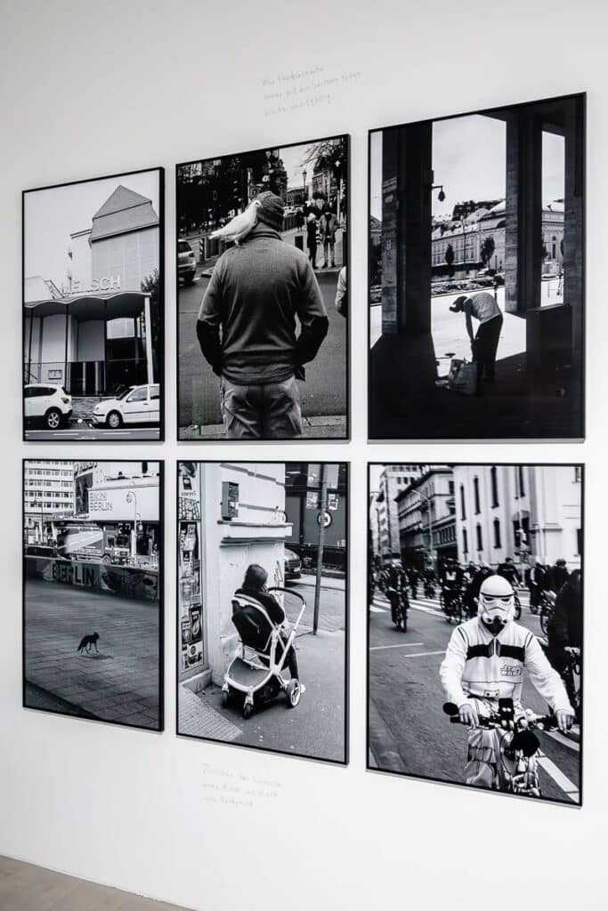 Sechs Schwarzweißfotografienwerden in zwei Reihen auf einer weißen Wand ausgestellt. Die Bilder zeigen urbane Straßenszenen mit Menschen und Stadtansichten. Handgeschriebener Text, Haikus, umgibt die Fotografien und liefert Kontext.