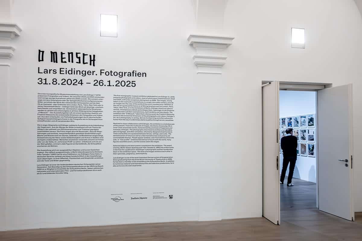 Eine Galerie mit weißen Wänden präsentiert die faszinierende Ausstellung „O Mensch“ von Lars Eidinger, die vom 31. August 2024 bis zum 26. Januar 2025 läuft. Im angrenzenden Raum betrachtet ein Besucher gerahmte Fotografien an der Wand. Der Haupttext erläutert Ausstellungsdetails und Künstlerinformationen.