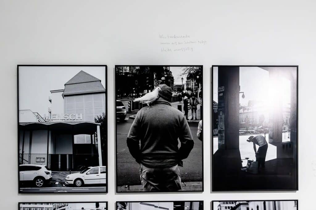Drei Schwarzweißfotos von Lars Eidinger hängen an einer weißen Wand im K21. Das linke Foto zeigt ein Gebäude mit dem Schriftzug „Mensch“. In der Mitte ist ein Mann mit einer Taube auf dem Kopf zu sehen. Auf dem rechten Foto ist jemand zu sehen, der sich auf dem Bürgersteig bückt, darüber ist ein handgeschriebener Text, ein Haiku vomnm Yoko Tawada.